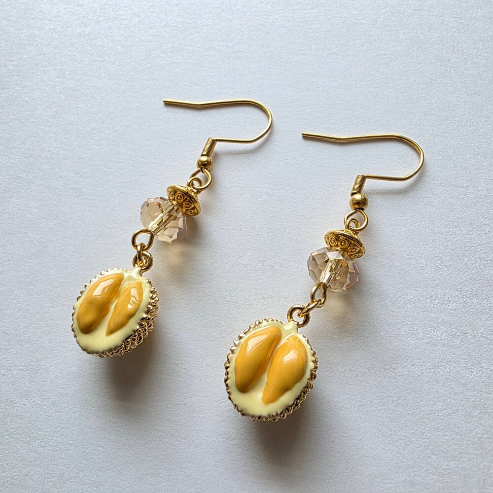 A pair of gold-tone dangle earrings featuring oval cream-colored charms with glossy yellow-orange details, suspended from clear faceted beads on a light gray background.