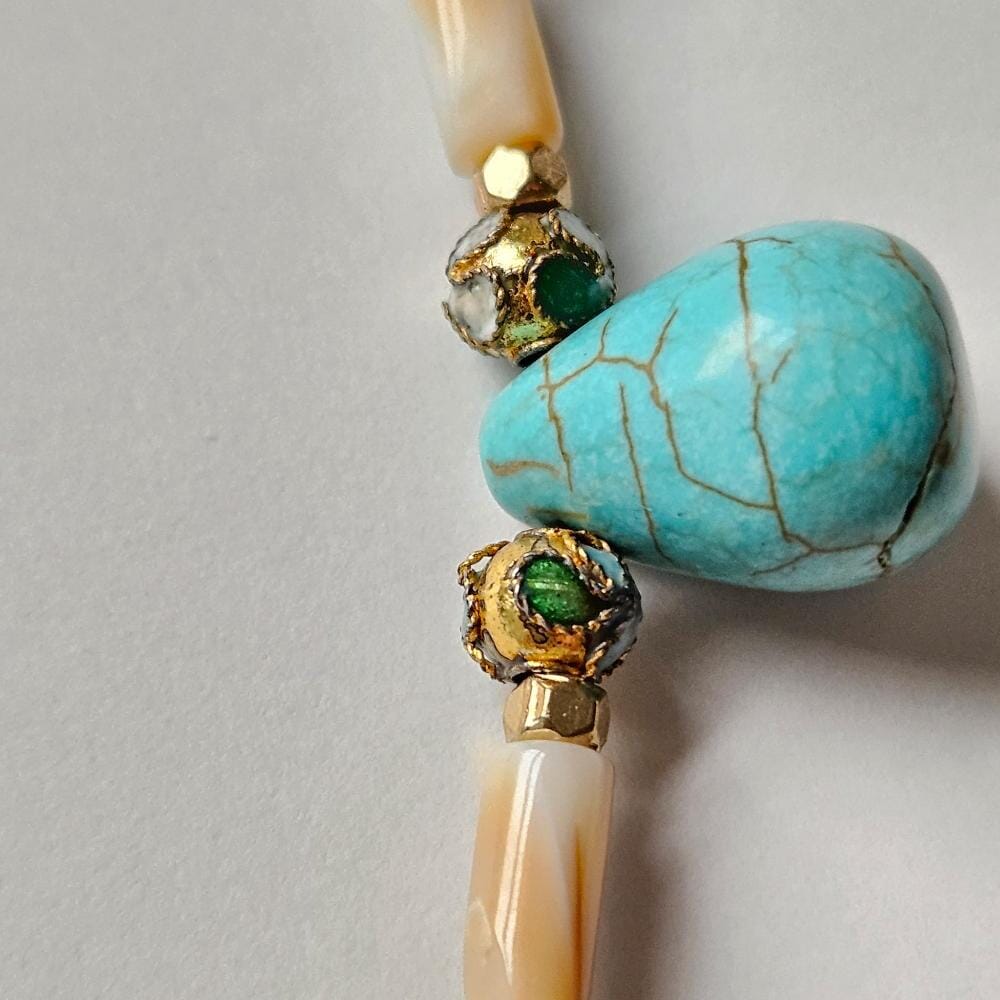 
                  
                    Close up detail of dyed turquoise colored howlite teardrop bead with cloisonne beads, gold colored metal faceted spacer beads, and mother-of-pearl tube beads.
                  
                